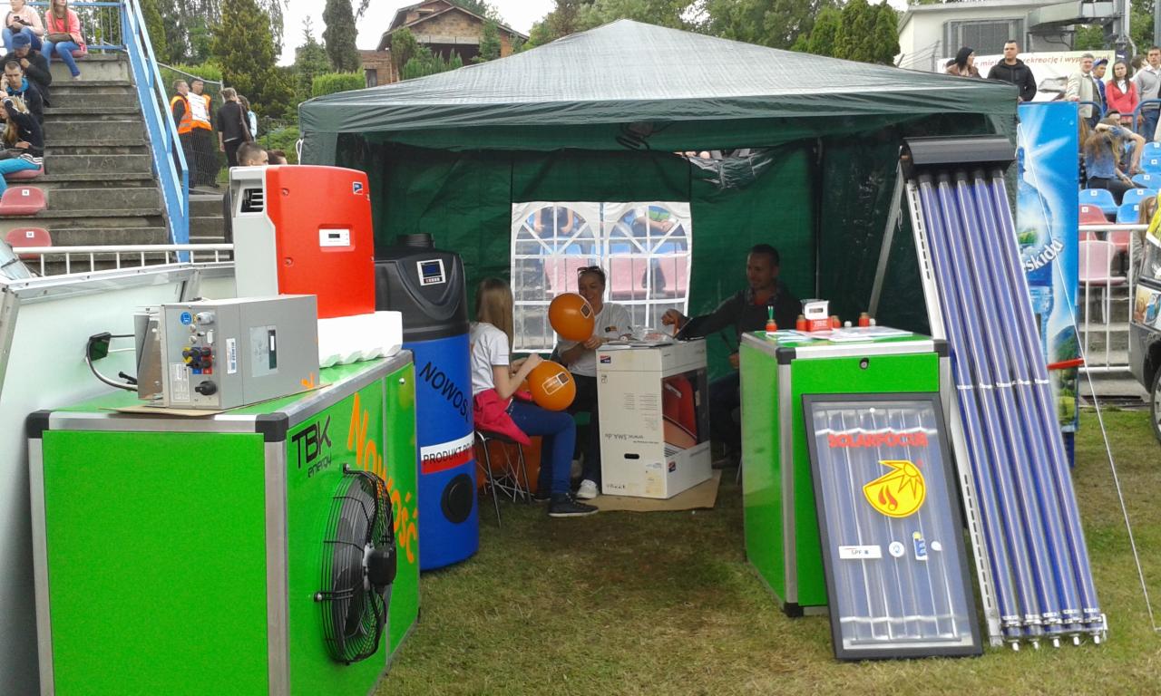 Ekspozycja systemów grzewczych i solarnych pod namiotem na zewnątrz, z osobami w tle. Widoczne kolektory słoneczne, pompy ciepła i inne komponenty instalacji.