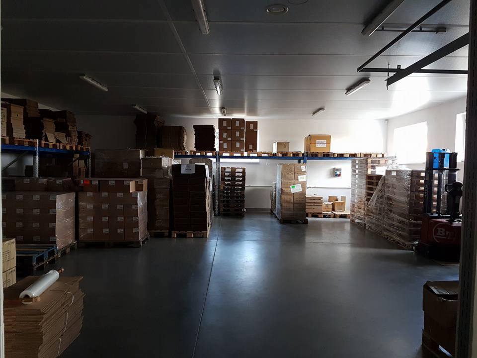 Zdjęcie przedstawia magazyn z kartonami ułożonymi na paletach i regałach, oświetlony światłem jarzeniowym, z widocznym wózkiem widłowym w tle, sugerujące etap przygotowania do wysyłki...