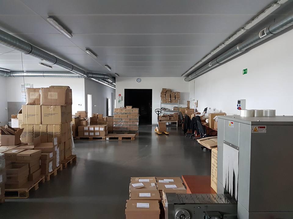 Wnętrze magazynu z paletami pełnymi kartonowych pudeł, widoczny wózek paletowy oraz maszyna pakująca z taśmociągiem.