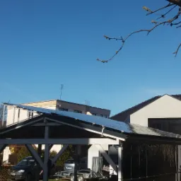 Kaskadowa instalacja paneli na wiacie garażowej, widoczny budynek mieszkalny w tle, błękitne niebo i gałąź drzewa w prawym górnym rogu.