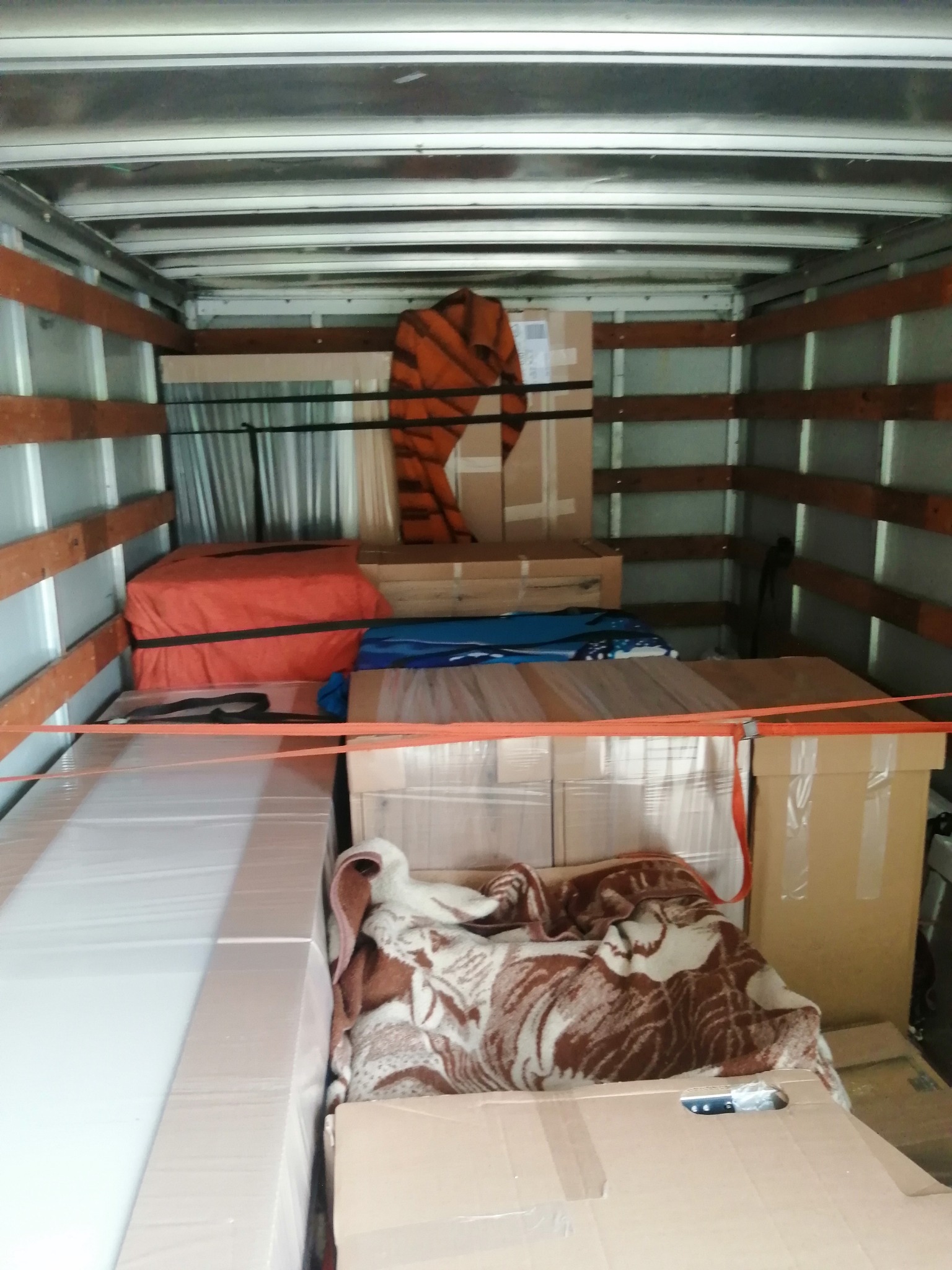 Wnętrze furgonetki załadowane kartonami, meblami owiniętymi folią i kocami, zabezpieczone pomarańczowymi pasami transportowymi.