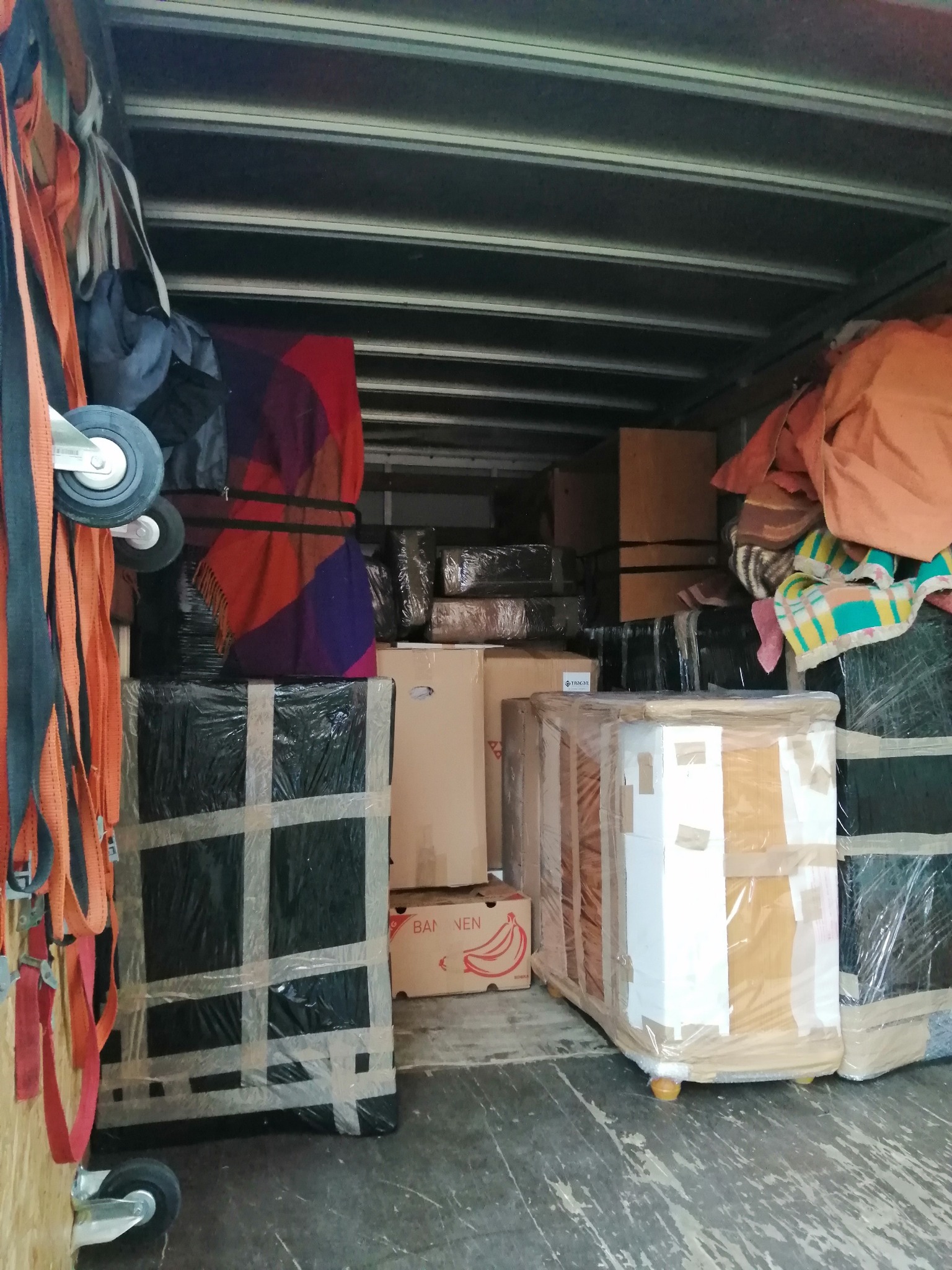 Wnętrze samochodu dostawczego wypełnione zapakowanymi meblami, kartonami i kocami, zabezpieczonymi folią i taśmą, widoczne pasy mocujące z kółkami na ścianie pojazdu.