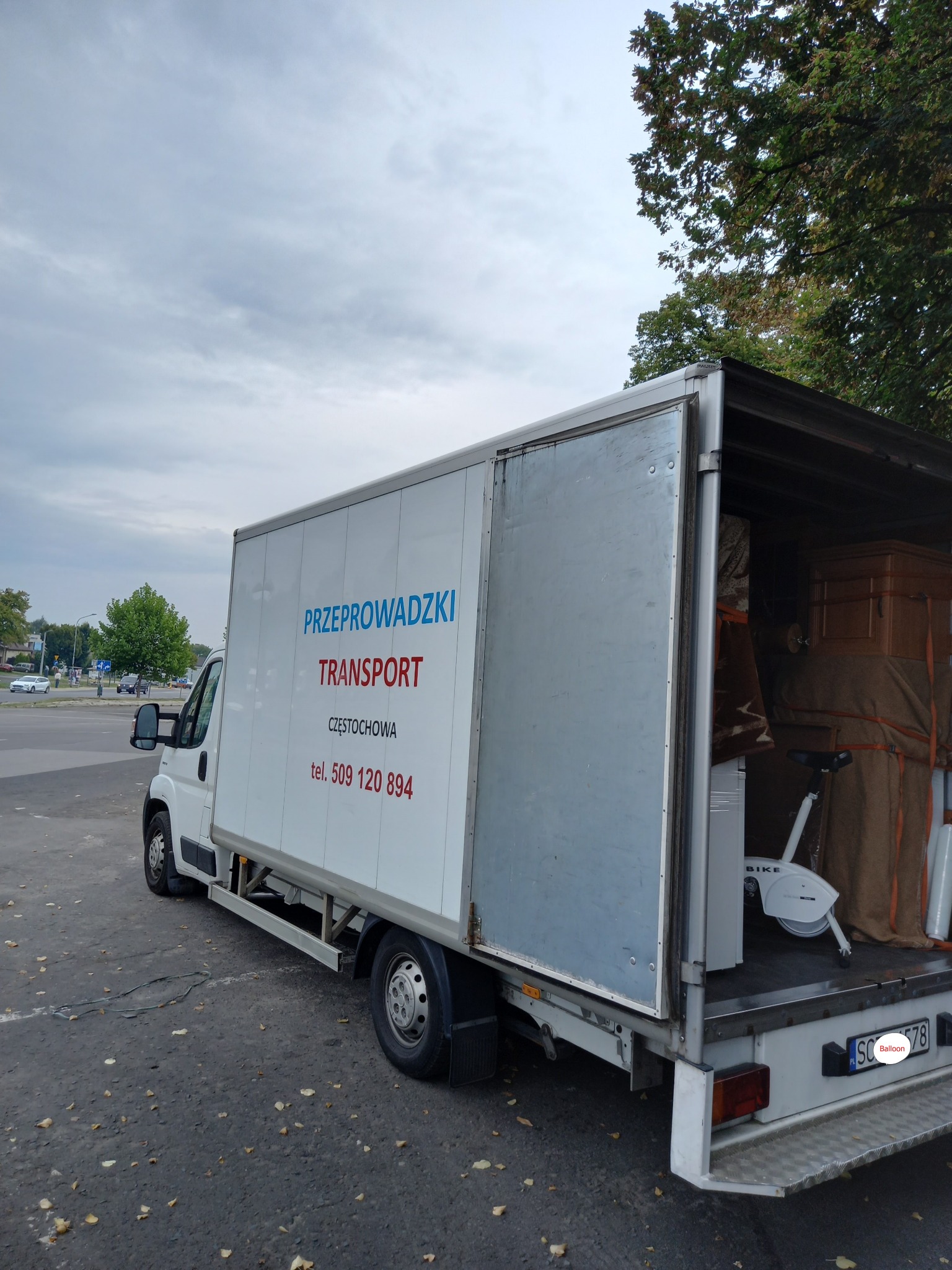 Częściowo otwarty biały samochód dostawczy z napisem 'Przeprowadzki Transport Częstochowa' załadowany meblami i sprzętem sportowym, w tym rowerem stacjonarnym.