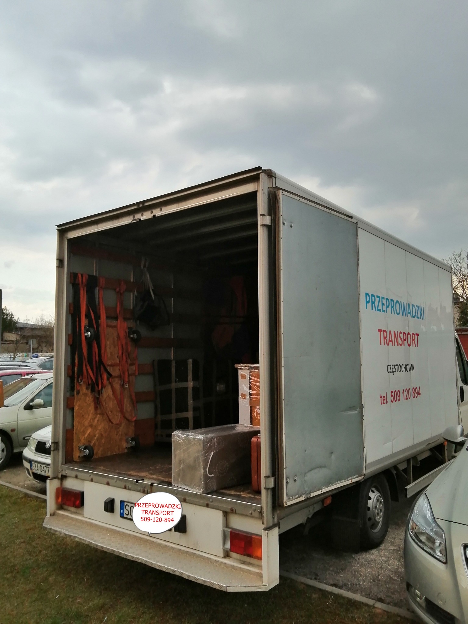 Otwarta paka samochodu dostawczego z widocznymi zabezpieczeniami ładunku i częścią przewożonych przedmiotów, na boku nadruk z nazwą firmy i numerem telefonu, na parkingu przed budynkiem.