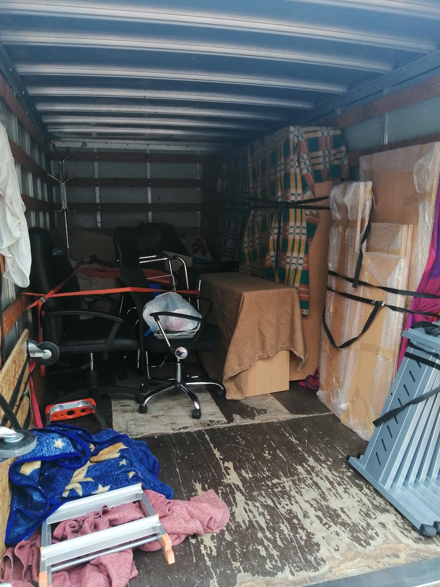 Wnętrze wypełnionej meblami i przedmiotami skrzyni ładunkowej, zabezpieczonymi pasami, widok na krzesła biurowe, stół przykryty brązowym materiałem, koce i drabiny.