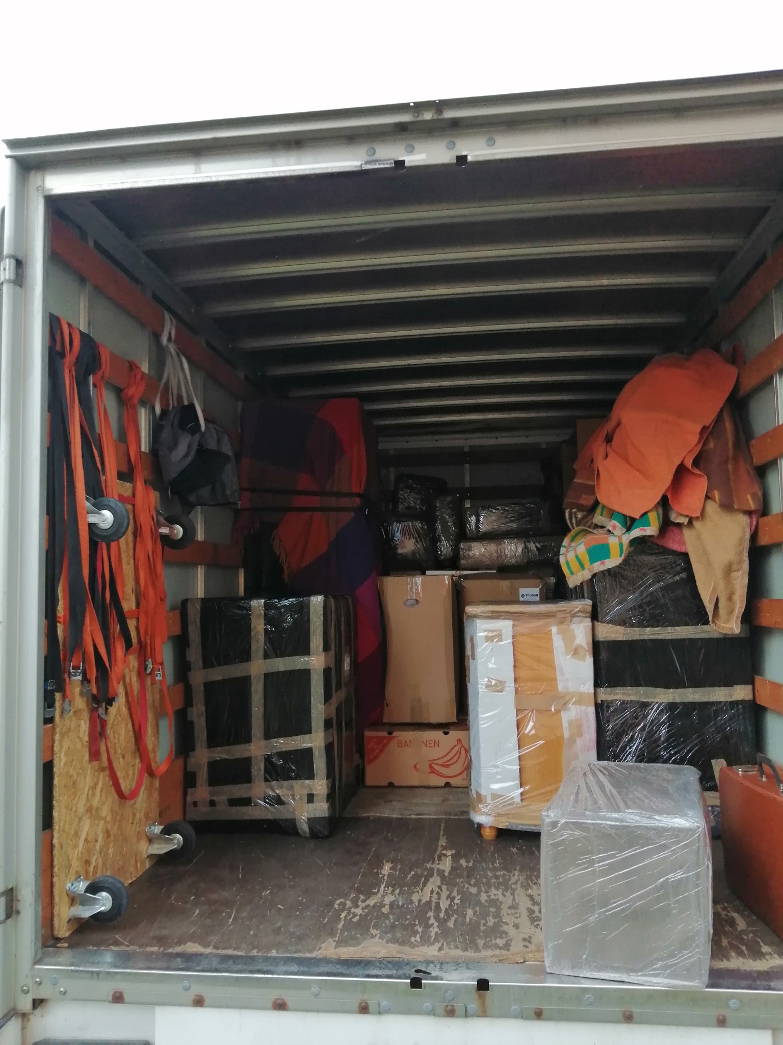 Wnętrze furgonetki załadowane kartonami i meblami zabezpieczonymi folią i taśmą, widoczne pasy mocujące i wózek transportowy.