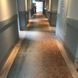 KERAJ-Podłogi - Długi korytarz hotelowy z nową wykładziną dywanową w odcieniach szarości i pomarańczy, widoczne panele podłogowe po bokach, w tle sylwetka osoby.