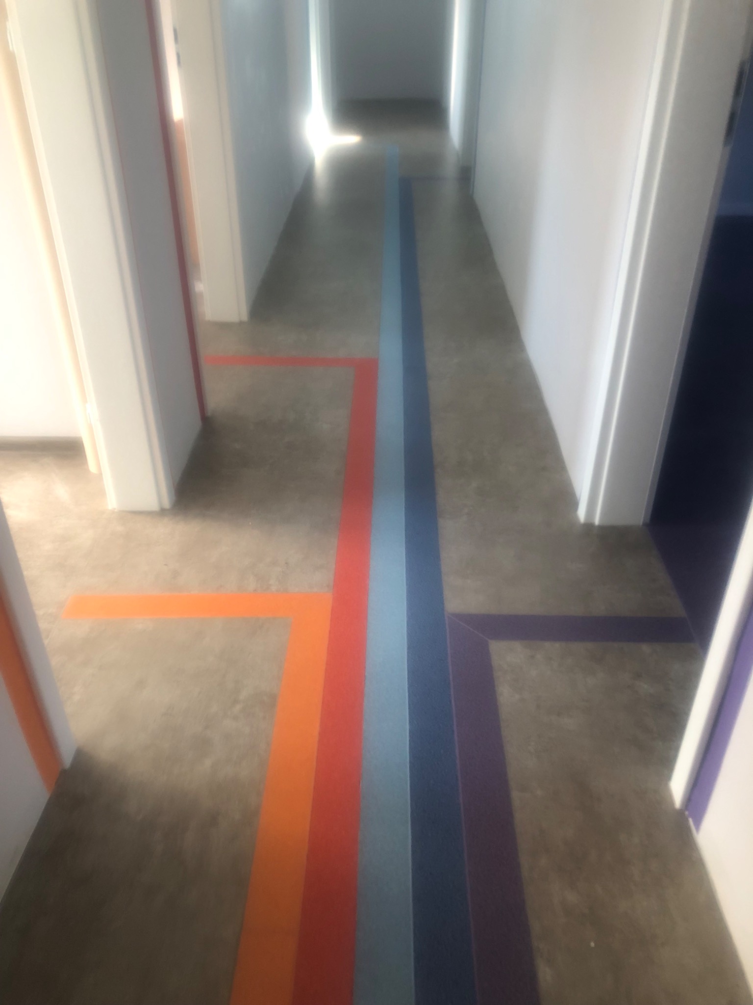 Długi korytarz z szarą podłogą, na której ułożono kolorowe pasy wykładziny w odcieniach pomarańczy, czerwieni, błękitu i fioletu, tworzące geometryczny wzór.