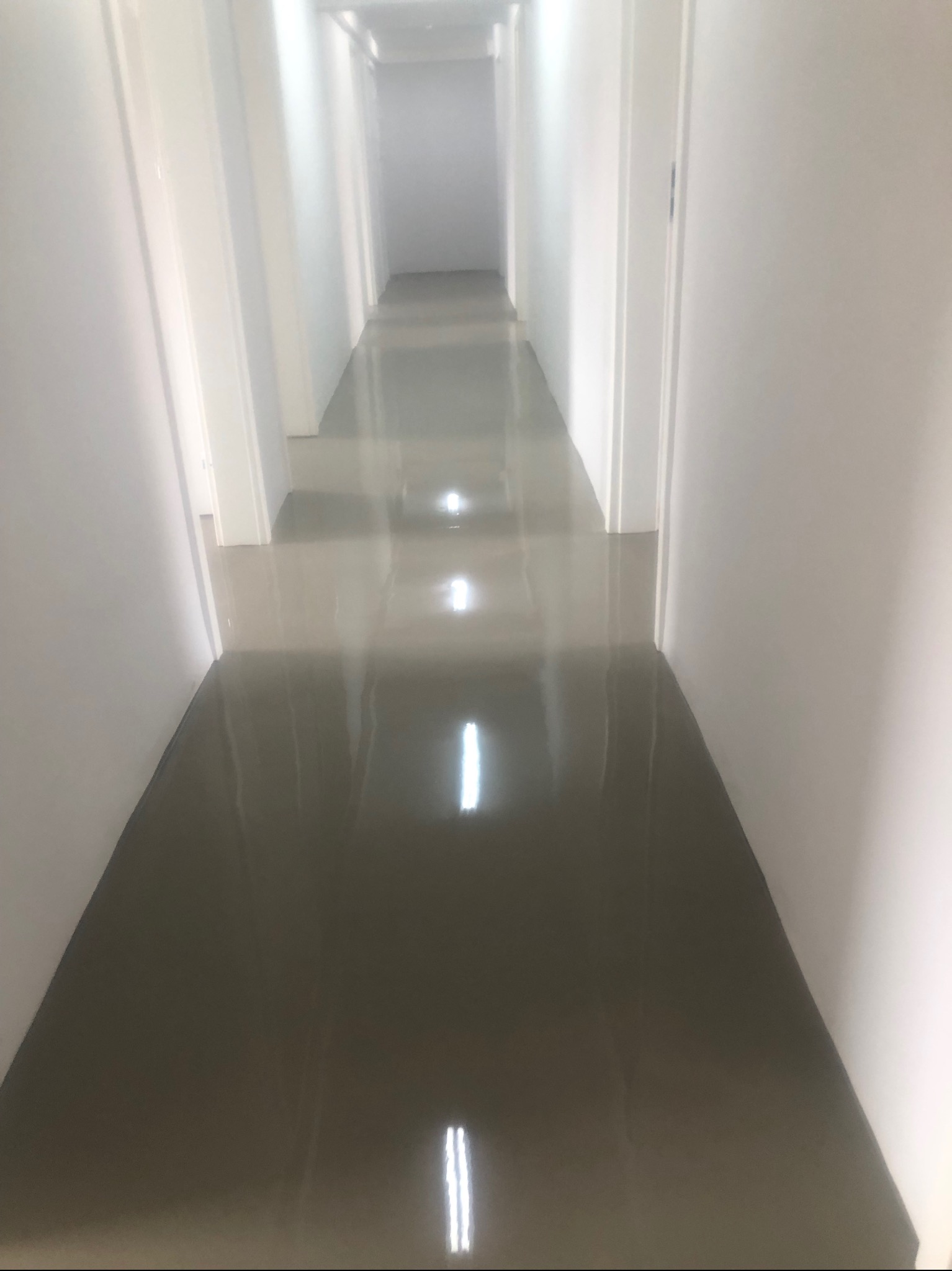 Długi korytarz z błyszczącą, gładką podłogą w odcieniach szarości, odbijającą światło, białe ściany i drzwi po obu stronach, perspektywa zbiegająca się w oddali.