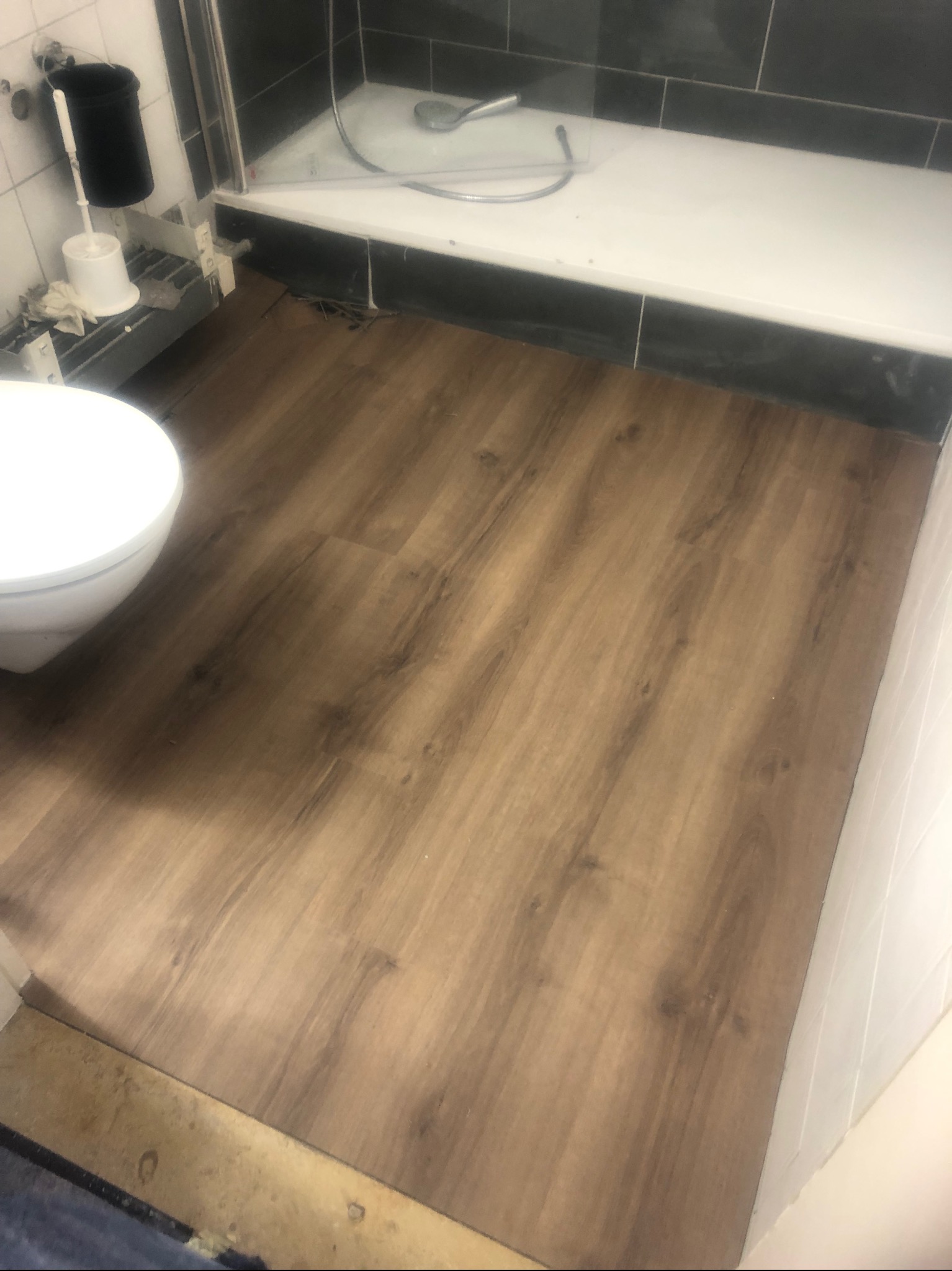 Ułożone panele podłogowe w łazience, widoczny fragment toalety i brodzika, jasnobrązowe panele z wyraźnym wzorem drewna.