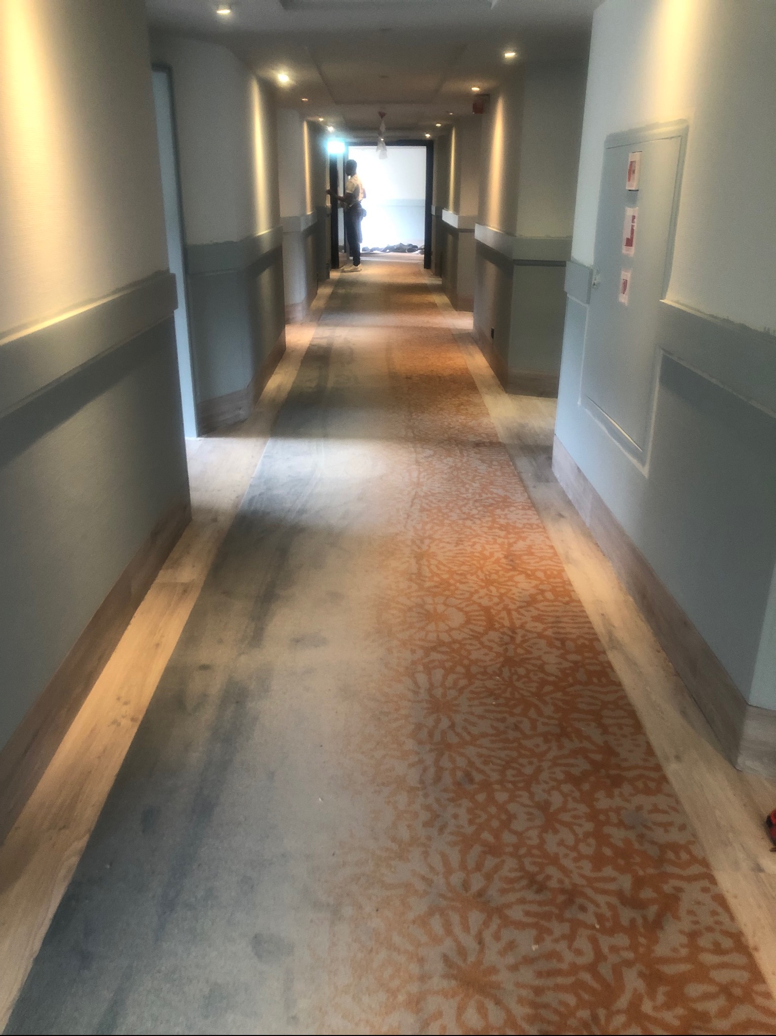 Długi korytarz hotelowy z nową wykładziną dywanową w odcieniach szarości i pomarańczy, widoczne panele podłogowe po bokach, w tle sylwetka osoby.