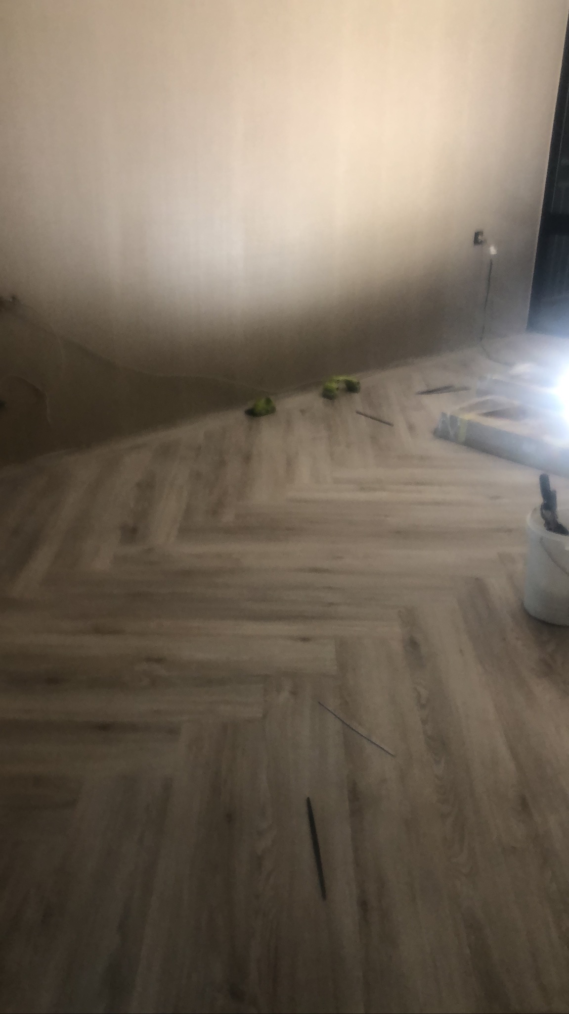 Ułożone panele podłogowe w jodełkę, widoczny biały pojemnik z narzędziami, zielone podkładki dystansowe oraz listwy przypodłogowe czekające na montaż.