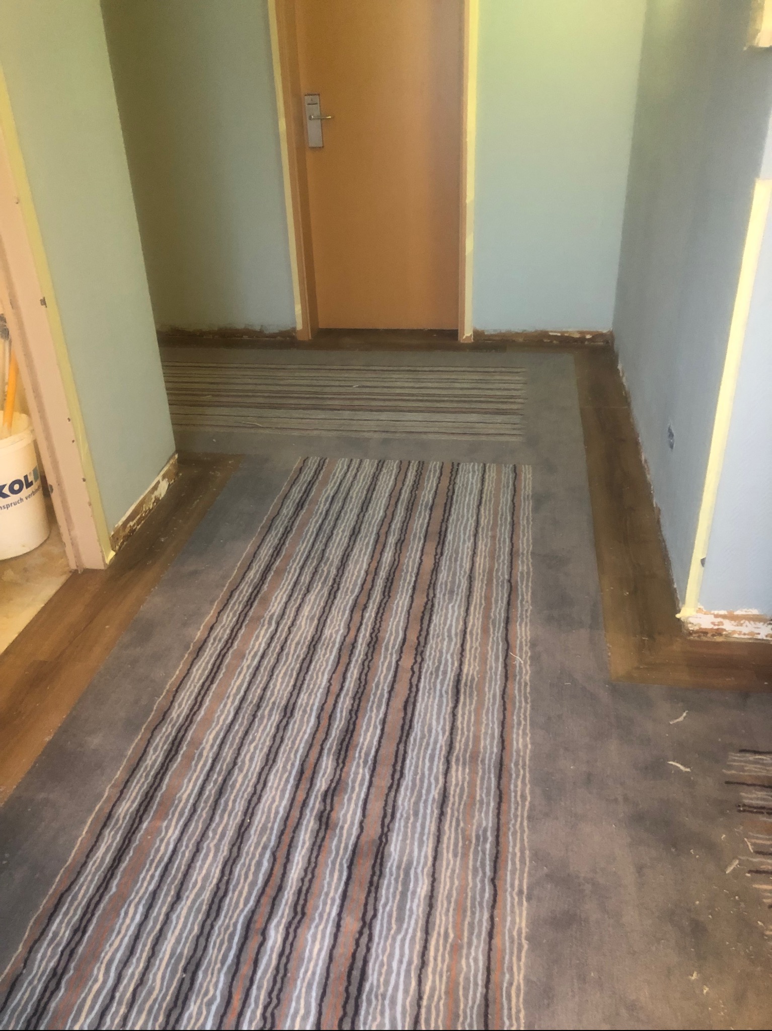 Korytarz w trakcie remontu z częściowo ułożoną wykładziną w paski i widocznymi śladami demontażu starej podłogi przy ścianach i futrynach drzwiowych.
