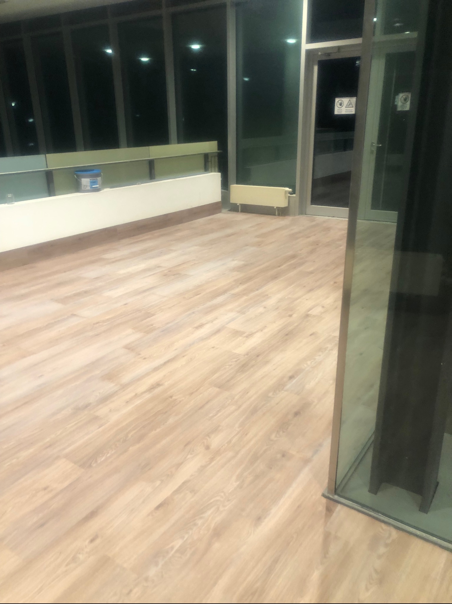 Jasne panele podłogowe ułożone w nowoczesnym biurze z dużymi oknami i widocznym pojemnikiem z klejem w tle.