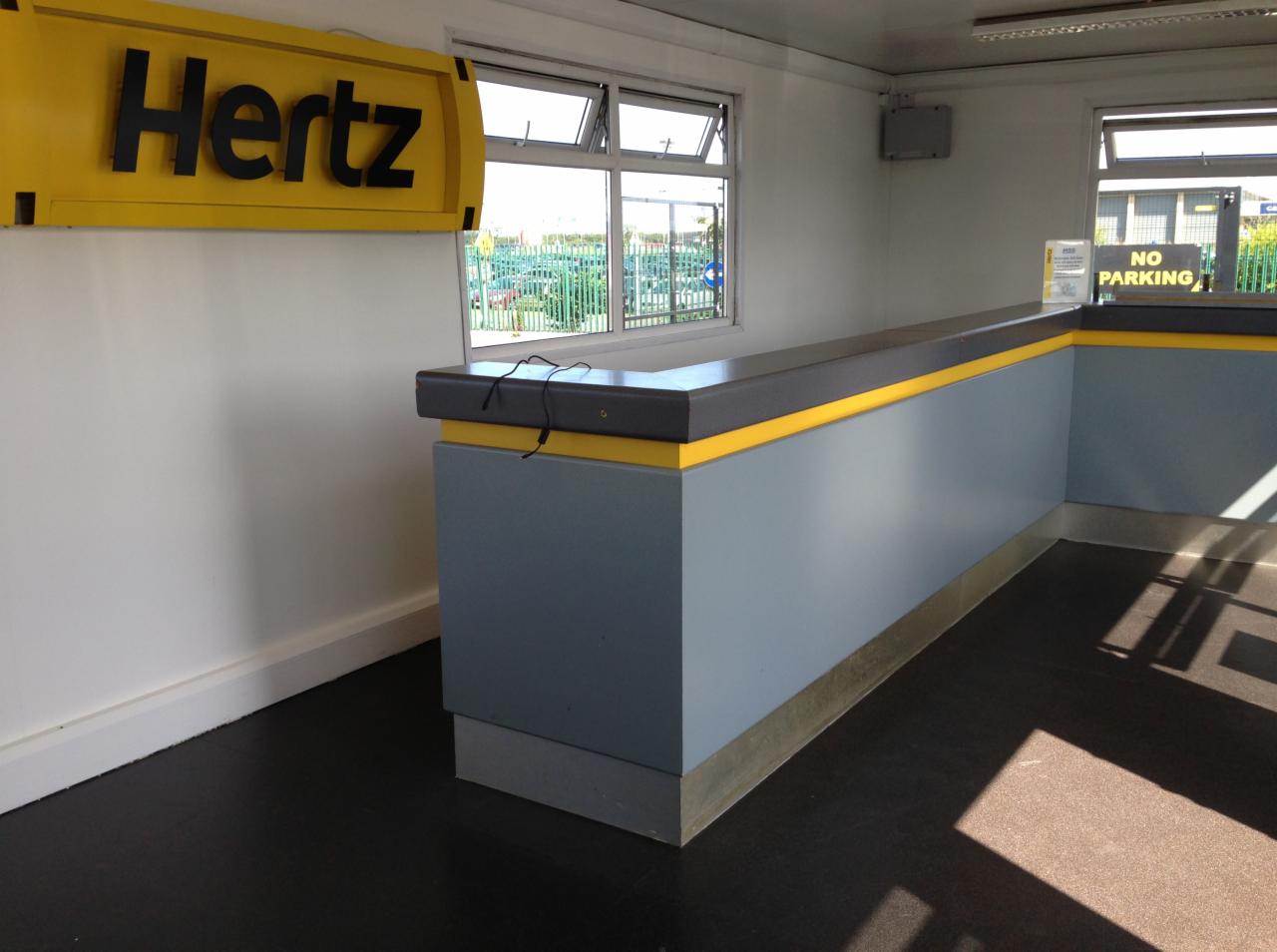 Wnętrze biura Hertz z czarną podłogą, szarą ladą z żółtym akcentem i oknami widocznymi na zewnątrz.