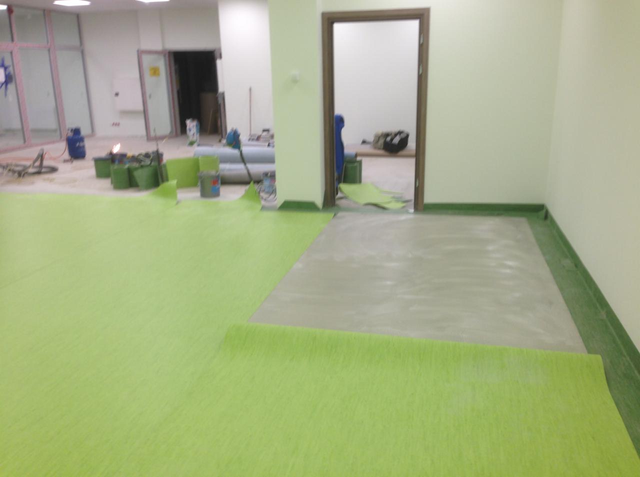 Proces montażu zielonej wykładziny podłogowej w jasnym pomieszczeniu; widoczne narzędzia, klej i częściowo położona wykładzina na przygotowanym podłożu betonowym.