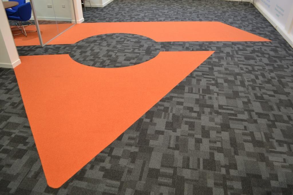 Wzorzysta podłoga biurowa z szarej wykładziny dywanowej z geometrycznym, pomarańczowym akcentem w kształcie fragmentu koła i trójkąta.