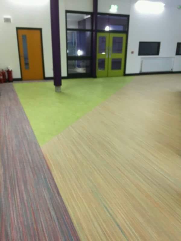 Widok na fragment podłogi w dużej sali z trzema różnymi typami nawierzchni: wykładzina w paski po lewej, jednolita zielona wykładzina w środku oraz panele imitujące drewno po prawej. W tle widoczne...