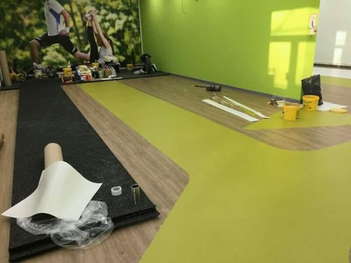 Wnętrze siłowni w trakcie montażu podłogi: fragment czarnej wykładziny gumowej, rolka z białym papierem ochronnym, puszki z klejem i zielona, wylana podłoga z żywicy epoksydowej w tle.