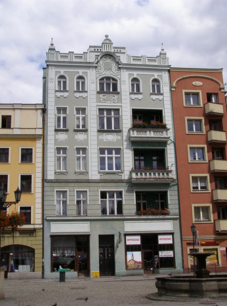 Fasada kamienicy w stylu secesyjnym z balkonami i oknami, obok budynek w kolorze terakoty. Na witrynach widnieją napisy 'WYPRZEDAŻ' i 'NA SPRZEDAŻ'.