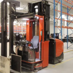 Pomarańczowo-czarny wózek widłowy elektryczny typu reach truck w magazynie z regałami paletowymi w tle.