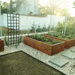 Ogród na dachu we Wrocławiu: podwyższone drewniane grządki z nasadzeniami, czarna kratka na pnącza, ścieżka z płyt betonowych na żwirowym podłożu, widok na ogrodzenie i budynki w tle, logo firmy...