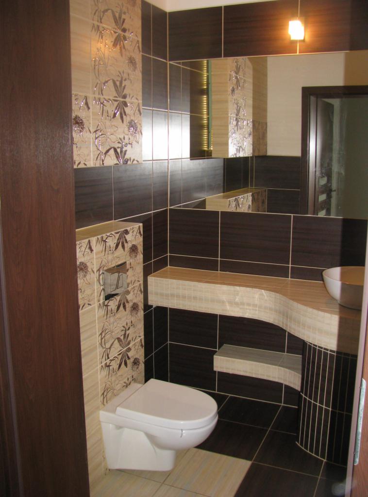 Wykończona łazienka z brązowo-beżowymi płytkami, toaleta podwieszana i umywalka na blacie. Elementy dekoracyjne z motywem kwiatowym. Lustro nad umywalką.