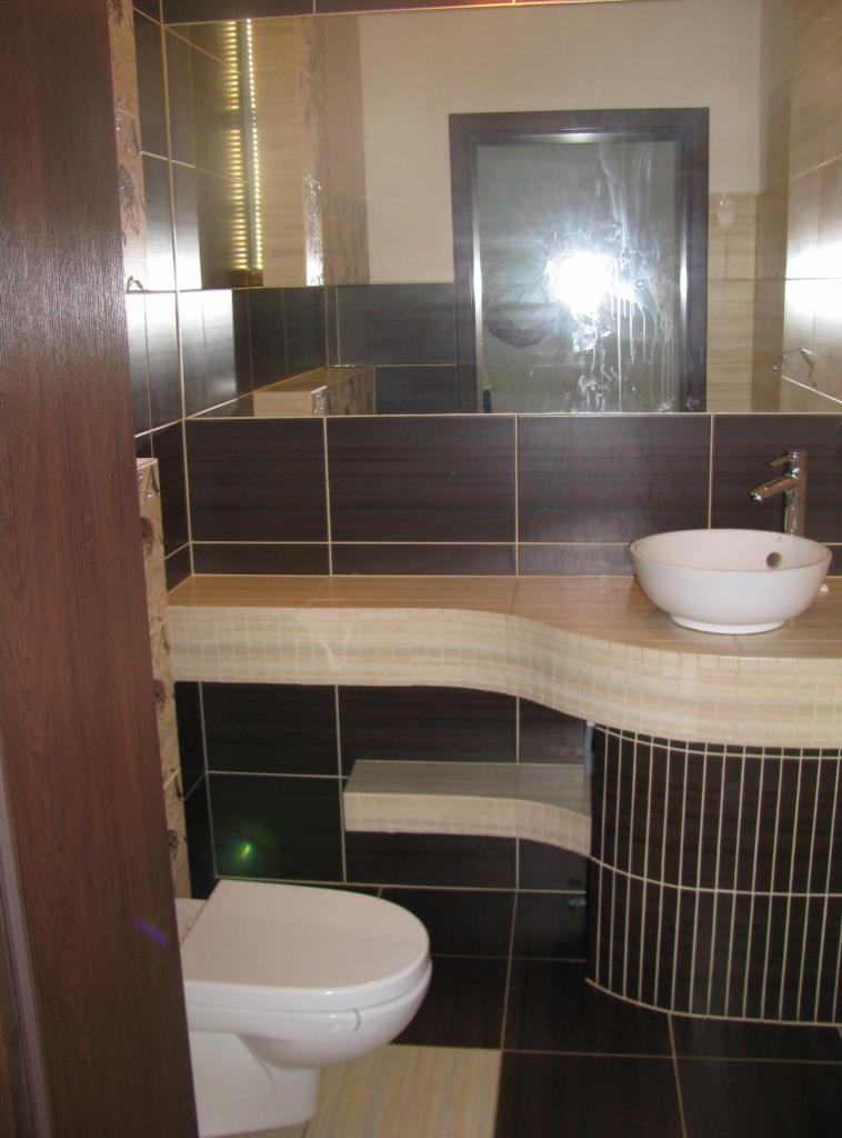 Wykończona łazienka z brązowymi i beżowymi płytkami. Umywalka nablatowa, lustro i toaleta. Widoczne elementy wykończenia i aranżacji wnętrza.