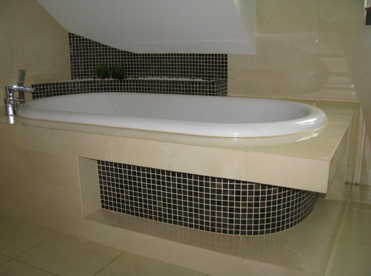 Nowoczesna wanna obudowana beżowymi płytkami, z mozaiką w czarno-białą kratkę. Bateria chromowana, w tle małe roślinki w doniczkach. Całość w jasnej tonacji.