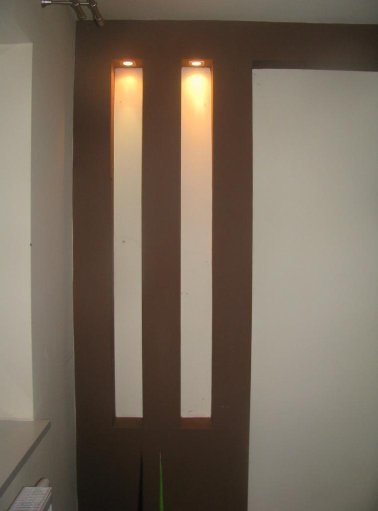 Nowoczesna ściana z dwoma podświetlanymi wnękami pomalowana w kolorze brązowym, obok biała ściana. Widoczne rury pod sufitem i fragment blatu.