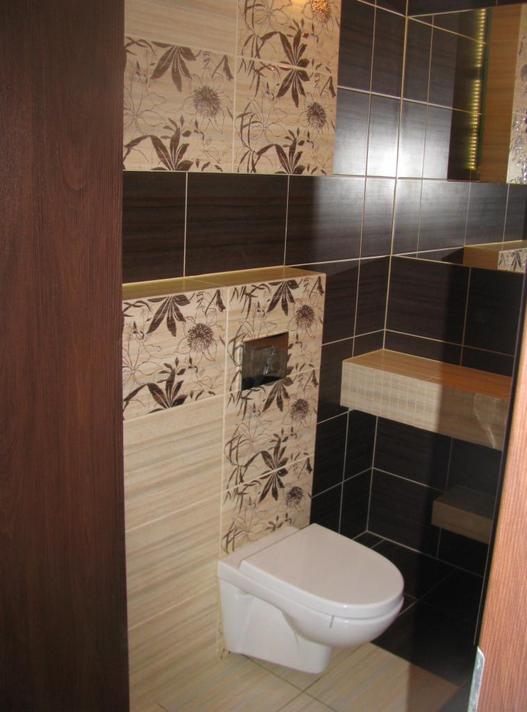 Wykończona łazienka z podwieszaną toaletą i dekoracyjnymi brązowo-beżowymi płytkami w kwiatowe wzory. Na ścianie widoczny chromowany przycisk spłuczki i półka.
