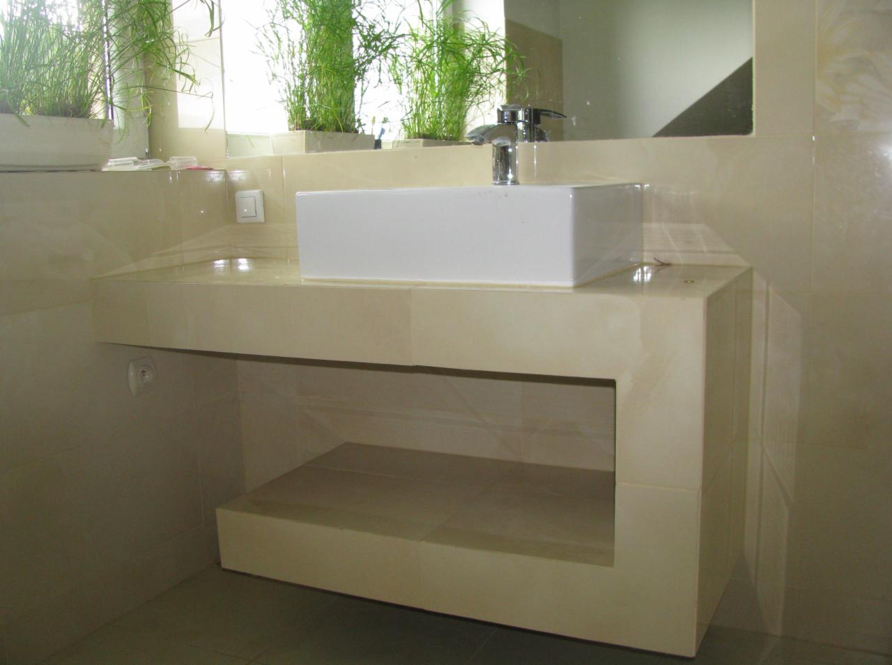 Beżowa łazienka z prostokątną umywalką nablatową i roślinami w tle. Umywalka osadzona na zabudowie z półką. Całość wykończona płytkami.