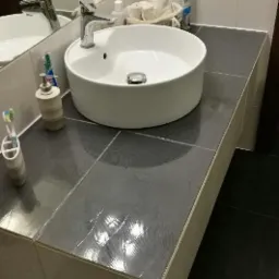 Umywalka nablatowa, okrągła, na szarym blacie z płytek w łazience z białymi ścianami, widoczne akcesoria łazienkowe i lustro.