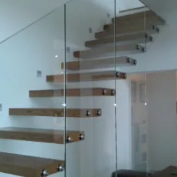 Nowoczesne schody dywanowe z drewnianymi stopniami i szklaną balustradą, mocowane do ściany. Widok z boku, minimalistyczny design, jasne wnętrze.