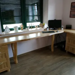 Długi, jasny, drewniany blat biurka z naturalną krawędzią, podparty na nogach imitujących gałęzie, z szufladami po bokach, ustawiony pod oknem z zielonymi żaluzjami, z komputerem i dekoracjami...