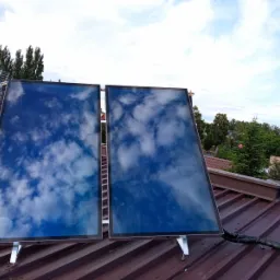 Dwa kolektory słoneczne na brązowym dachu, odbijające błękitne niebo z chmurami, w tle dom z anteną i drzewa.