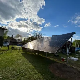 Instalacja paneli słonecznych na trawiastym terenie przydomowym, widoczne akcesoria montażowe i drabina, błękitne niebo z promieniami słońca odbijającymi się w panelach.