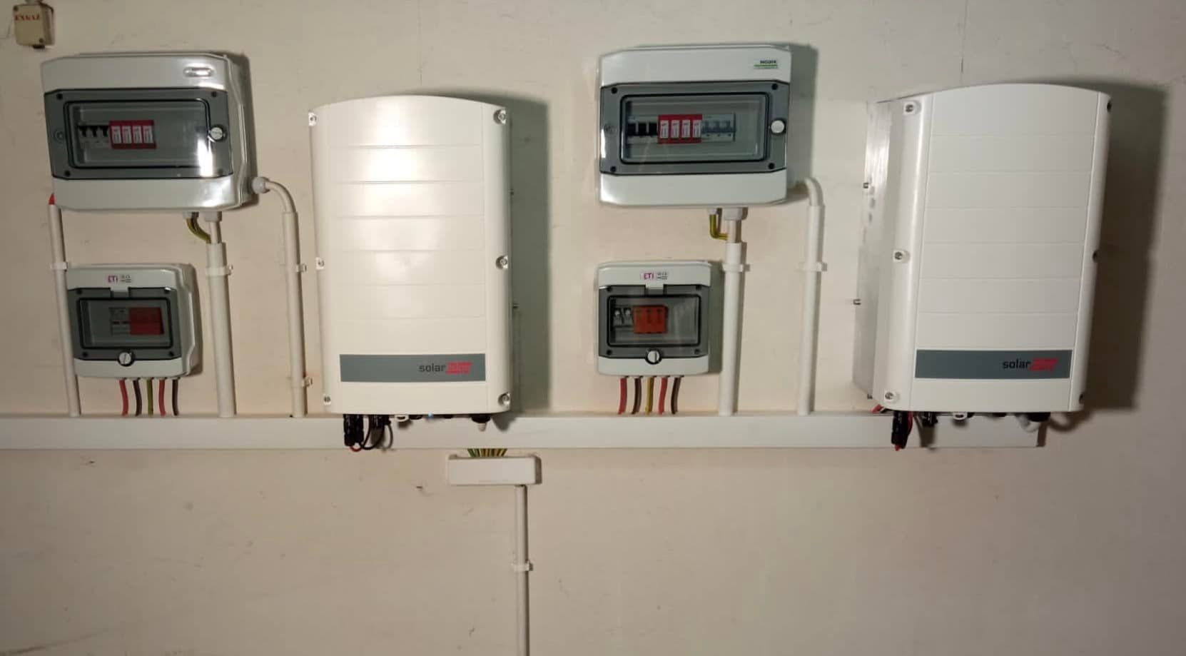 Dwa białe falowniki SolarEdge i skrzynki bezpiecznikowe zamontowane na białej ścianie wewnątrz budynku.
