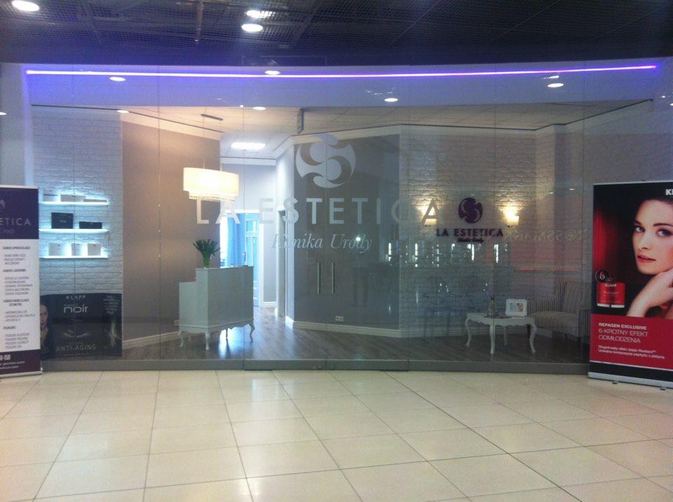 Eleganckie wejście do salonu kosmetycznego 'La Estetica' z przeszklonymi drzwiami, logo na szkle, recepcją i banerem reklamowym z wizerunkiem kobiety.
