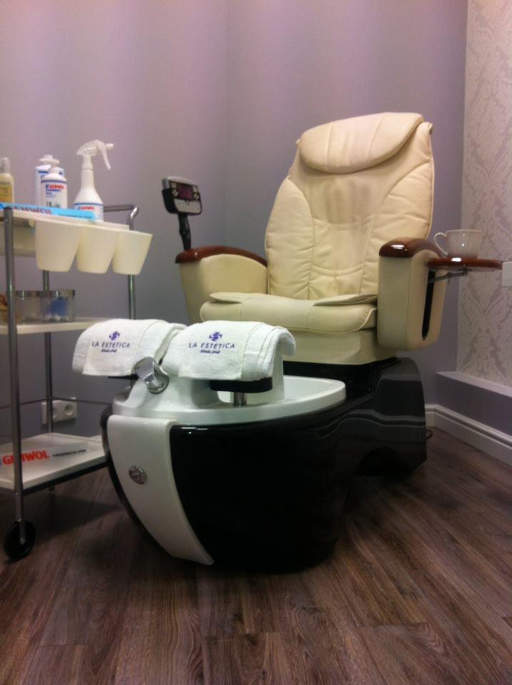 Fotel do pedicure z podnóżkiem i wózkiem kosmetycznym. Na podnóżku leżą białe ręczniki z logo 'La Estetica'. Obok fotela stoi filiżanka. Całość w salonie kosmetycznym.