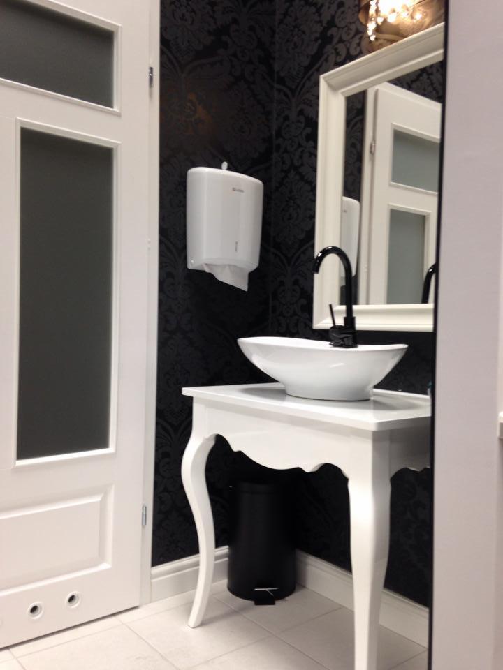 Elegancka toaleta w salonie: biała umywalka na ozdobnym stoliku, czarna tapeta w ornamenty, lustro i dozownik ręczników. Estetyczne wykończenie.