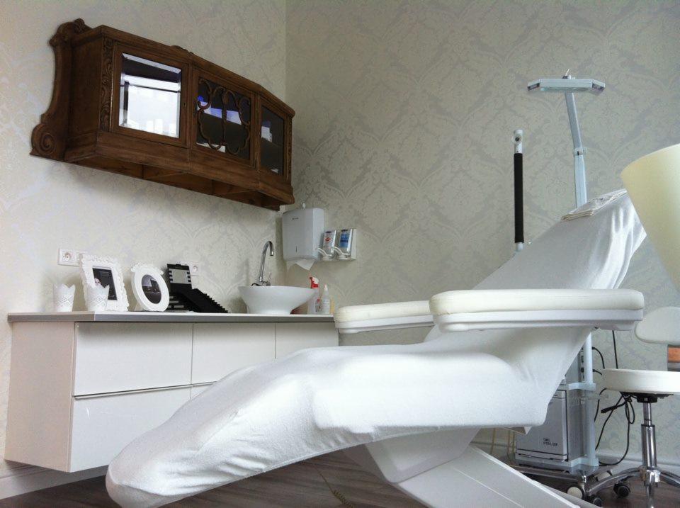 Jasne wnętrze gabinetu kosmetycznego z fotelem zabiegowym przykrytym białym pokrowcem i umywalką na tle ściany w delikatny wzór.