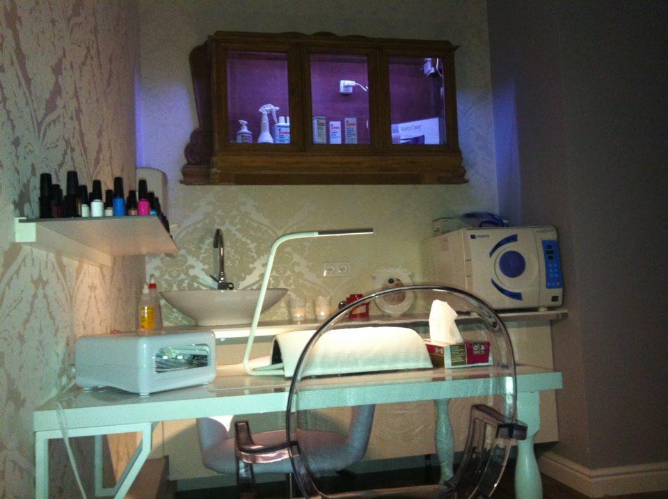 Stanowisko do manicure z lampą UV, pochłaniaczem pyłu i licznymi lakierami na półce. Nad blatem szafka z kosmetykami i umywalka w salonie kosmetycznym.