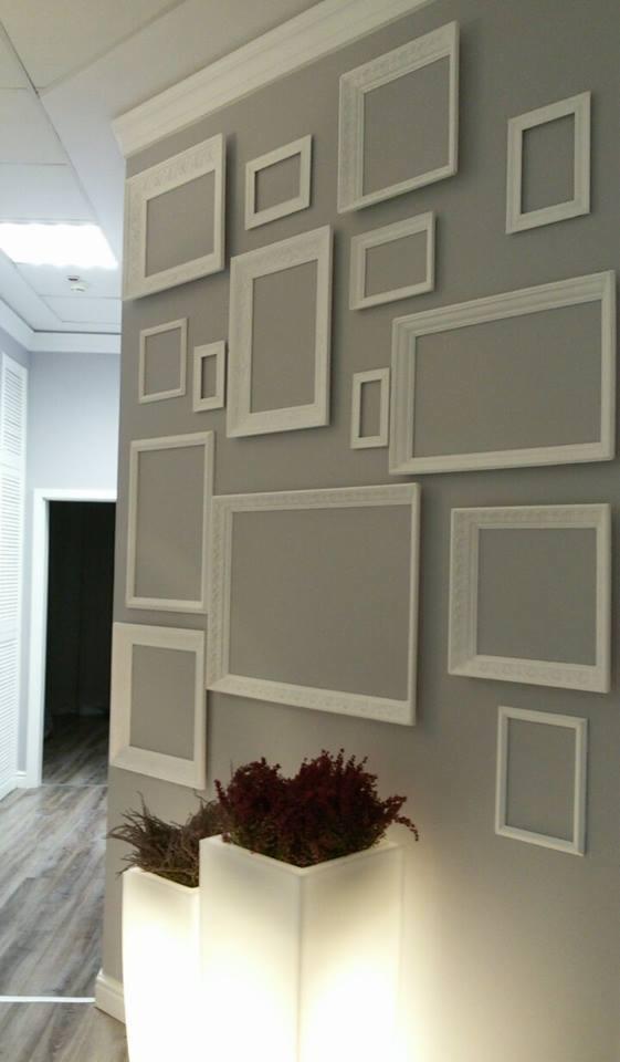 Ściana w salonie kosmetycznym zdobiona białymi ramkami o różnych rozmiarach, podświetlane donice z roślinami. Widoczny fragment podłogi i drzwi.