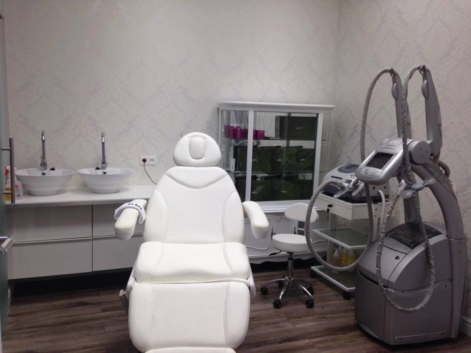 Nowoczesny gabinet kosmetyczny: biały fotel zabiegowy, umywalki, szafka i sprzęt do modelowania sylwetki w jasnym wnętrzu z wzorzystą tapetą.