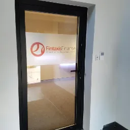 Szklane drzwi wejściowe do biura z naklejonym logo i nazwą 'Fintaxis Finanse. Departament Wsparcia Biznesu' w kolorze czerwonym, widok na wnętrze biura z jasnymi ścianami i podłogą.