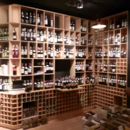 Regał na wino z jasnego drewna, wypełniony butelkami wina, tworzący ekspozycję w sklepie. Podłoga drewniana, kartony u dołu. Ciemne tło.