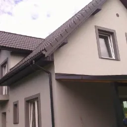 Dwukondygnacyjny dom z szarą elewacją, brązowym dachem z dachówki i szarymi obramowaniami okien, widoczny z perspektywy narożnej.