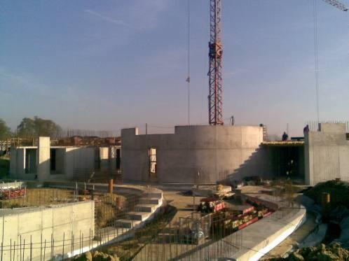 Surowy betonowy szkielet budynku w trakcie budowy, z widocznym żurawiem w tle i schodami prowadzącymi do górnej części konstrukcji.