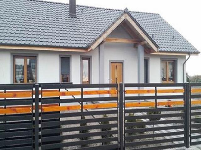 Nowoczesny dom jednorodzinny z szarą dachówką i drewnianymi oknami, ogrodzony metalowym płotem z drewnianymi elementami w kolorze miodowym.